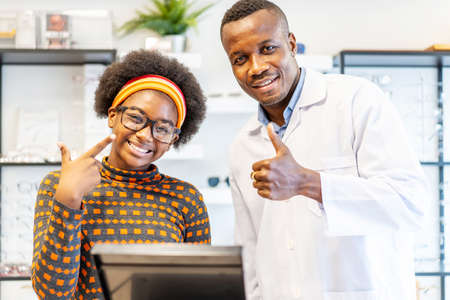 Professional ophthalmologist doctor african american man helping young woman african american client to choose a spectacles is successful. The girl is glad to have new glasses.の写真素材