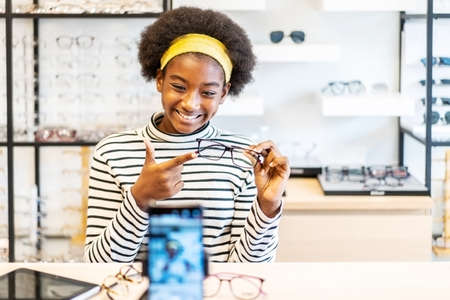 Young woman african american entrepreneur is fashion vlogger show glasses to cellphone and smiling selling it online live streaming in eyeglasses shopの写真素材