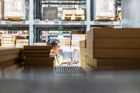 Asian female worker in safety vest sitting and working with computer laptop in storage warehouse. people, warehouse and industry conceptの写真素材