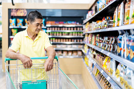 Asian senior man shopping trolley choosing other products in supermarketの写真素材