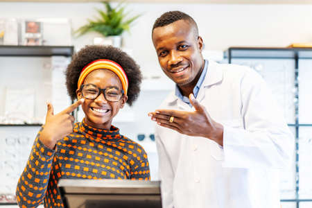 Professional ophthalmologist doctor african american man helping young woman african american client to choose a spectacles is successful. The girl is glad to have new glasses.の写真素材