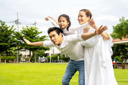 Portrait of asian happy family of three having fun together in park, little daughter sitting on father back and mother smile walking and having fun moments good time in summer park at homeの写真素材