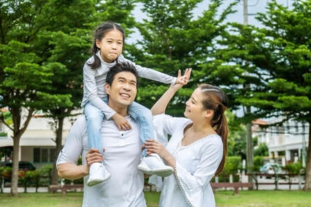 Portrait of asian happy family of three having fun together in park,  little daughter sitting on father back and mother smile walking and having fun moments good time in summer park at homeの写真素材
