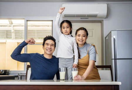 Happy family having fun in the kitchen. Asian Father, mother and little daughter spending time together and having breakfast drinking and hold glasses of milk at tableの写真素材