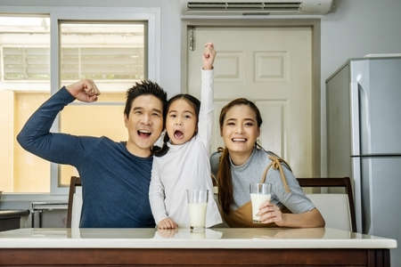 Happy family having fun in the kitchen. Asian Father, mother and little daughter spending time together and having breakfast drinking and hold glasses of milk at tableの写真素材