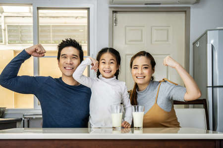Happy family having fun in the kitchen. Asian Father, mother and little daughter spending time together and having breakfast drinking and hold glasses of milk at tableの写真素材