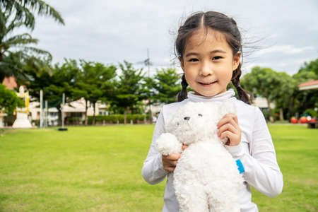 Happy asian little child girl standing in the grass and playing with her favorite teddy bear toy outdoors in summer parkの写真素材