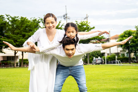 Portrait of asian happy family of three having fun together in park, little daughter sitting on father back and mother smile walking and having fun moments good time in summer park at homeの写真素材