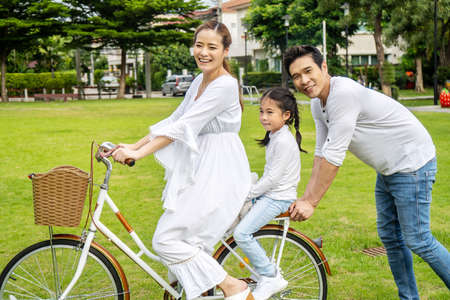 Happy family with bicycle outdoors on summer day, Asian father and mother with daughter enjoying riding a bike outdoors in a park.の写真素材