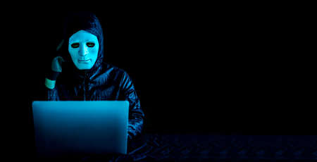 Hacker in white mask and hoodie using a laptop to browse the net and serious face thinking about...