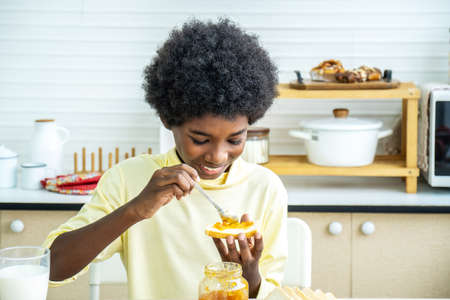 Cute african american boy spreading jam on bread and eating in kitchen, student enjoys breakfast or...
