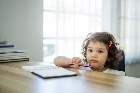 Cute little girl sit at desk at home doing homework, reading, writing and painting. Children paint. Kids draw. Preschooler with books at home. Preschoolers learn to write and read. Creative toddlerの写真素材