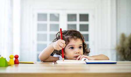 Cute little girl sit at desk at home doing homework, reading, writing and painting. Children paint. Kids draw. Preschooler with books at home. Preschoolers learn to write and read. Creative toddlerの写真素材