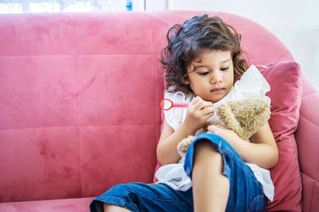A little cute girl enjoy playing toy injection syringes toy on the teddy bear on the sofa in the home, education time at homeの写真素材