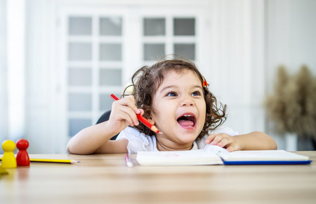 Funny little girl sit at desk at home doing homework, reading , writing and painting. Children paint. Kids draw. Preschooler with books at home. Preschoolers learn to write and read. Creative toddlerの写真素材