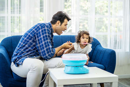 Hand father rolling cotton candy in candy floss machine and eat cotton candy together with daughter on sofa at new houseの写真素材