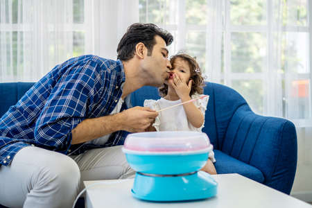 Caucasian family young father kissing his little daughter at mouth after eat cotton candy together on sofa at new houseの写真素材