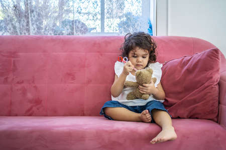 A little cute girl enjoy playing toy injection syringes toy on the teddy bear on the sofa in the home, education time at homeの写真素材