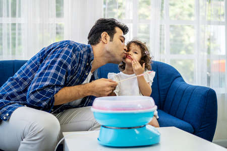 Caucasian family young father kissing his little daughter at mouth after eat cotton candy together on sofa at new houseの写真素材