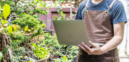 Man gardener working on laptop on some project surrounded by plants. use a laptop to monitor temperature control, and record leaf growth quality in the tree, Home gardening, freelance.の写真素材