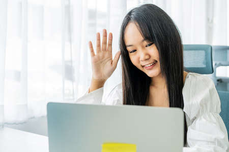 Smiling asian young female in headphones wave hand to laptop screen greet teacher tutor at lesson consultation. Happy young woman say hi hello to friend speaking from university by video callの写真素材