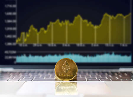 A golden ethereum bitcoin is place on the keyboard in front of a screen of stock or cryptocurrency market candles or charts or graphs with a laptop, cryptocurrency trading and investment conceptの写真素材