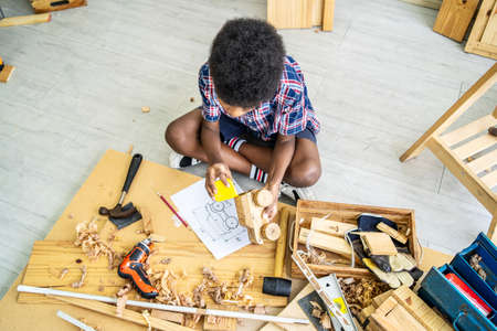 Cute little boy as a construction worker working with wood sitting at floor and taking measurements making wooden model, playing with toolsの写真素材