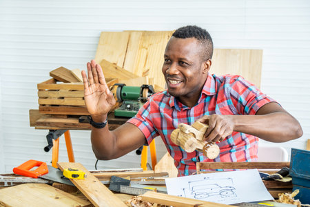 Young african american male carpenter sitting at table showing model car wooden and drawing paper through tablet while video online chatting with customer or teaching online in factoryの写真素材