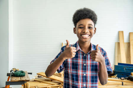 Smiling African-American boy carpenter standing with giving thumbs up as sign of success in a carpentry workshop.の写真素材