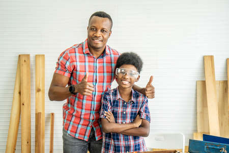 family, carpentry, woodwork, Portrait of cheerful african american father and little son making thumbs up at workshop, with diverse working tools laying on itの写真素材