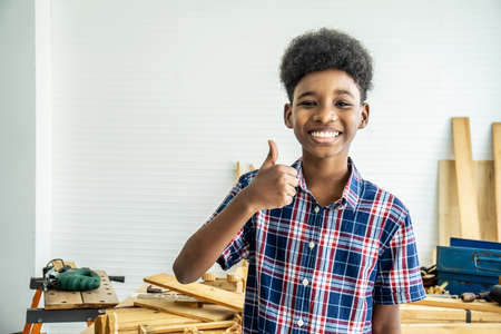 Smiling African-American boy carpenter standing with giving thumbs up as sign of success in a carpentry workshop.の写真素材
