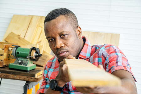 Young carpenter african american man looking and choosing wood and using sandpaper to rub wooden plank at workshop table in carpenter wood factoryの写真素材