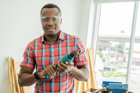 Work in the carpentry workshop. African american happy male holds a screwdriver in his hand. human in a woodworking workshop. Builder at the workplace. Carpenter poses in the workplace.の写真素材