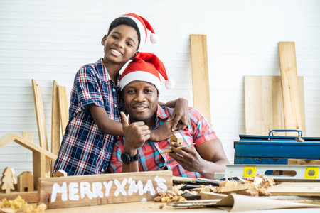 Christmas happy child and father . Cheerful african american son carpenter embracing his father while leaning at the wooden table with diverse working tools laying on itの写真素材