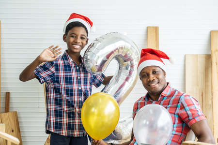 Christmas happy child and father. Cheerful african american son carpenter in santa hats having fun smiling at camera waving and saying hello to you with father, enjoying spending time togetherの写真素材