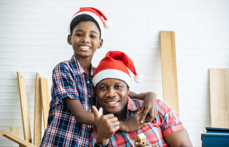 Christmas happy child and father . Cheerful african american son carpenter embracing his father while leaning at the wooden table with diverse working tools laying on itの写真素材