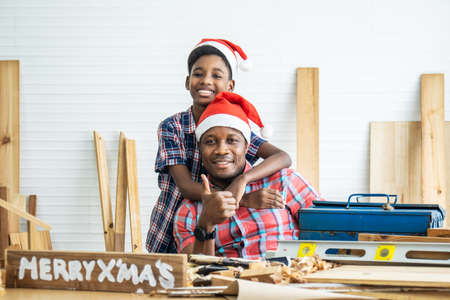 Christmas happy child and father . Cheerful african american son carpenter embracing his father while leaning at the wooden table with diverse working tools laying on itの写真素材