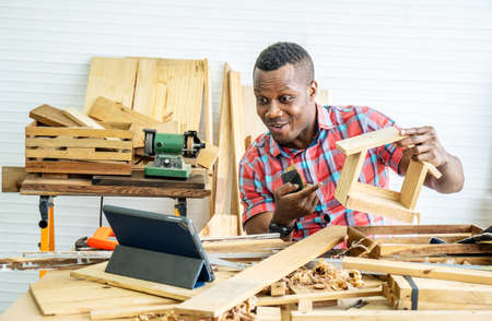 Young african american male carpenter sitting at table showing wooden items through tablet while video online chatting with customer or teaching online in factoryの写真素材