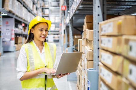 Asian engineer woman check stock details on laptop computer for checking boxes with logistics on shelves with goods background in warehouse, logistic and business exportの写真素材
