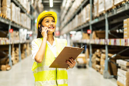 Asian woman in helmet and uniform talks on mobile phone discuss amount of stock product inventory on shelf at distribution warehouse factory. logistic business shipping and delivery serviceの写真素材