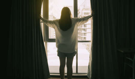 Overjoyed young beautiful woman standing by bedroom and opening curtains look in window distance...