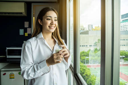 Young asian woman looking out window enjoying fresh new day feeling rested and drinking glass of milk at hotel.の写真素材