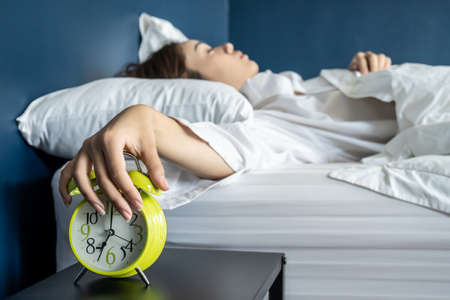 Close up sleepy woman stretching hand to ringing alarm willing turn it off, pressing button, woken by signal, lying under blanket in cozy bed, Early wake up, not getting enough sleep conceptの写真素材