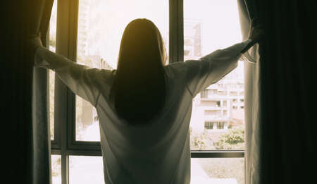 Overjoyed young beautiful woman standing by bedroom and opening curtains look in window distance meet welcome new day. Smiling female feel excited about life career opportunities or perspectives.の写真素材