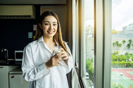 Young asian woman looking out window enjoying fresh new day feeling rested and drinking glass of milk at hotel.の写真素材