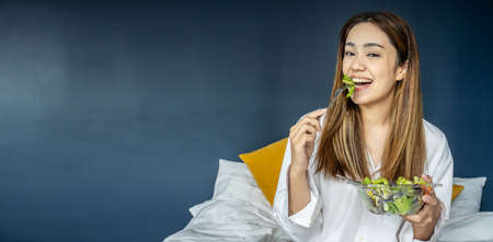 Gorgeous young woman sitting on bed eating vegetable salad, Balanced raw food diet. Positive girl having vegan lunch, taking care of her wellbeingの写真素材