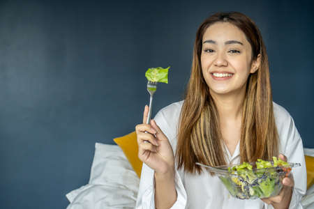 Gorgeous young woman sitting on bed eating vegetable salad, Balanced raw food diet. Positive girl having vegan lunch, taking care of her wellbeingの写真素材