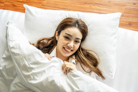 Cute lovely young woman lying in bed and under white blanket, looking at camera with eyes full of joy. Attractive girl having fun in bedroom.の写真素材