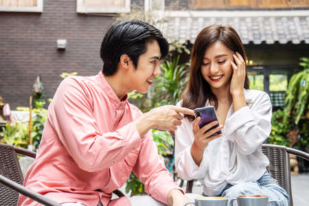 Happy Young asian woman sitting with boyfriend at cafe table, discussing over on smartphone, Young...