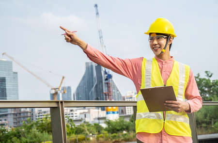 Asian male site contractor engineer with hard hat holding clipboard file and pen pointing at construction site, crane with golden sunlight at the background.の写真素材